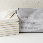 A stack of white organic cotton CloudCloth face cleansing cloths for makeup removal with a grey paper-wrapped package in the background, labelled with product information.